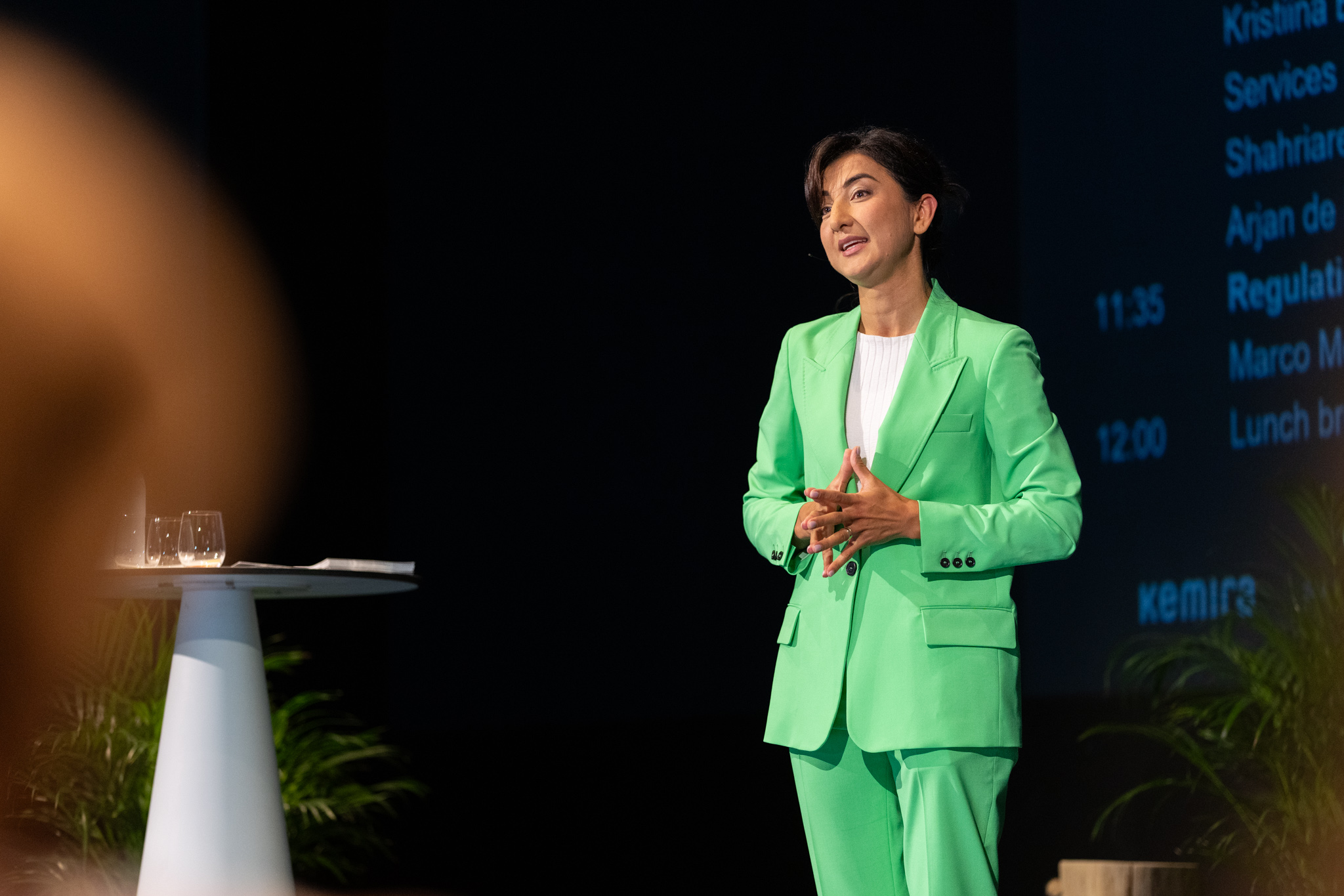 Kamilla standing on stage in a green suit hosting talk.